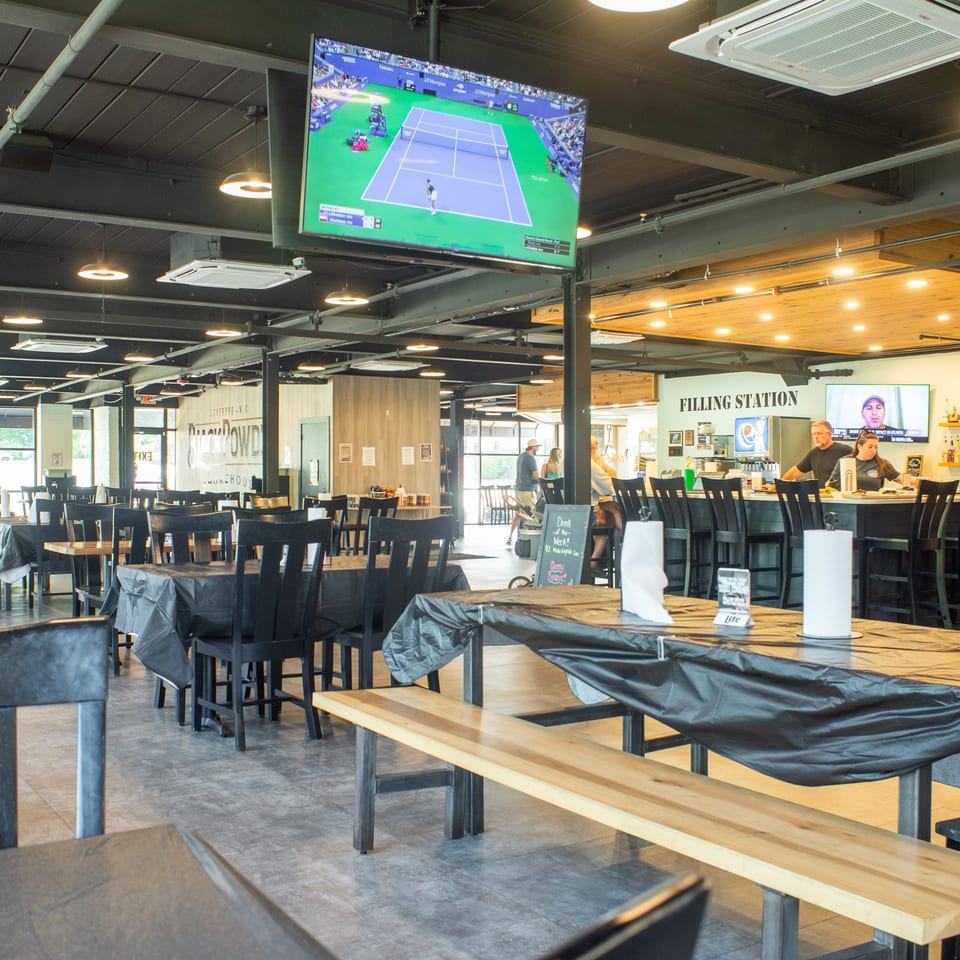 Spacious sports bar with communal tables, televisions showing tennis and baseball, a busy bar counter, and exposed industrial ceilings.
