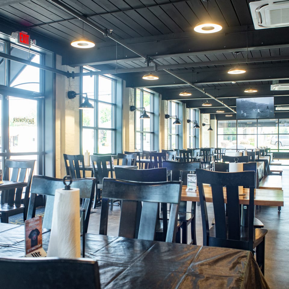 Spacious restaurant dining room with dark wood tables, black chairs, industrial lighting, and large windows.