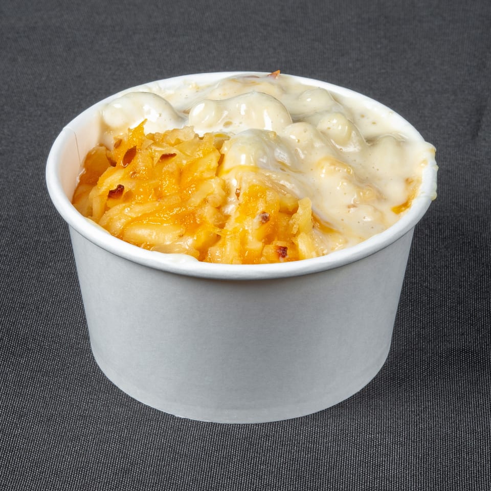 Paper cup filled with creamy macaroni and cheese topped with melted shredded cheddar on a dark textured surface.