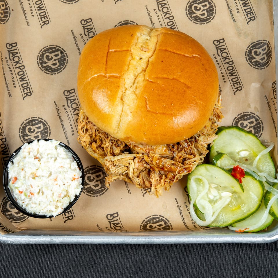 Pulled pork sandwich with barbecue sauce, coleslaw, and sliced cucumber salad on Black Powder Smokehouse serving paper.