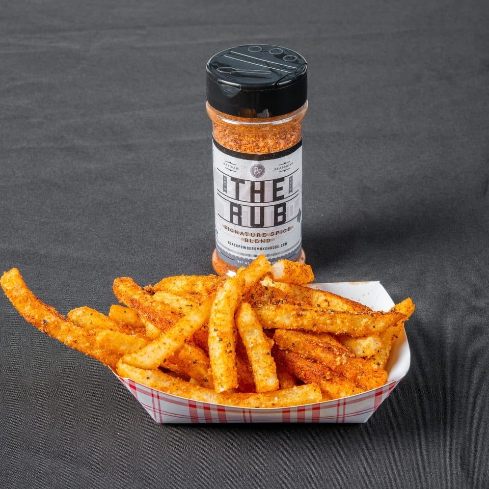 Seasoned French fries in a red-checkered tray sit before a bottle of The Rub signature spice blend.
