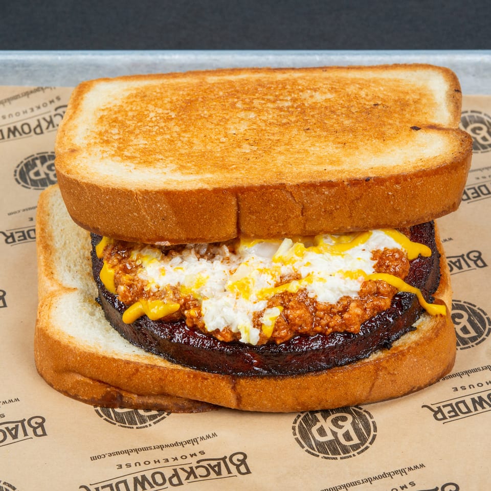 Close-up of a toasted sandwich layered with a dark beef patty, melted cheddar, fried egg, and saucy meat filling.
