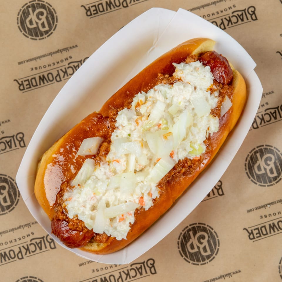 Chili dog topped with creamy coleslaw and diced onions in a toasted bun, served in a paper tray on branded butcher paper.