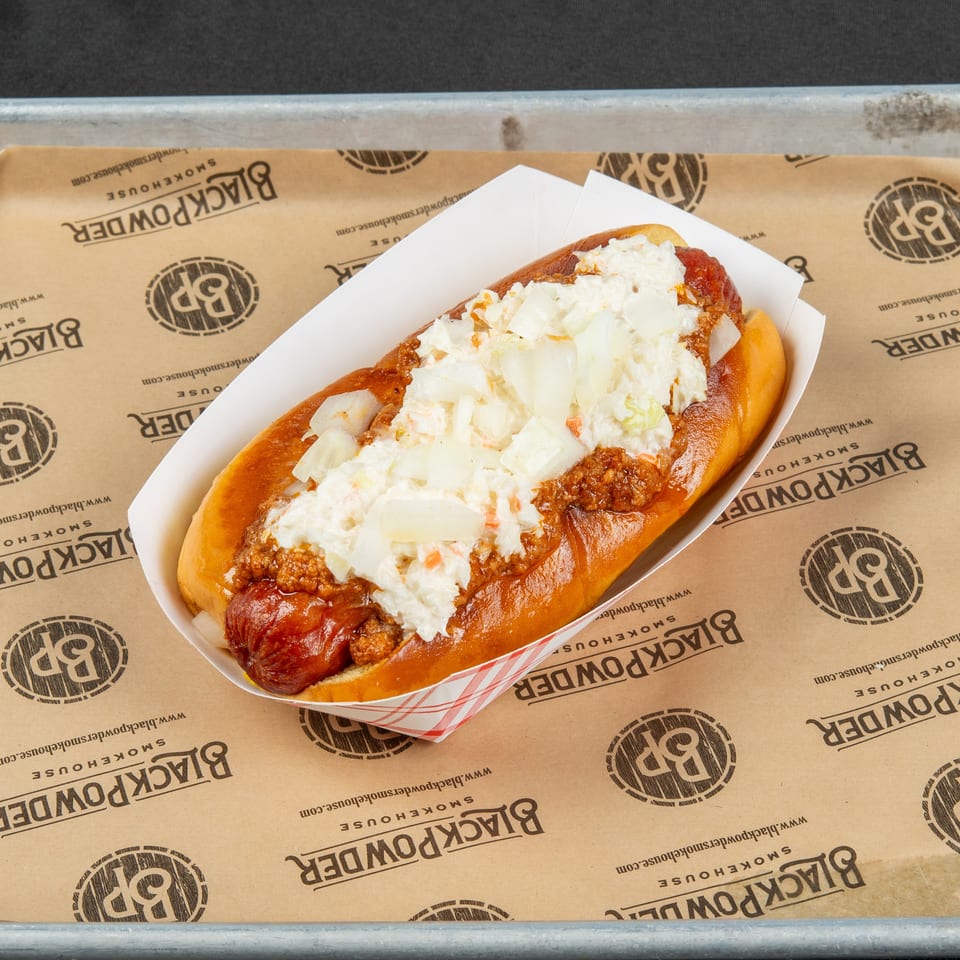 Chili dog topped with creamy coleslaw and diced onions in a paper tray on Black Powder Smokehouse-branded paper.
