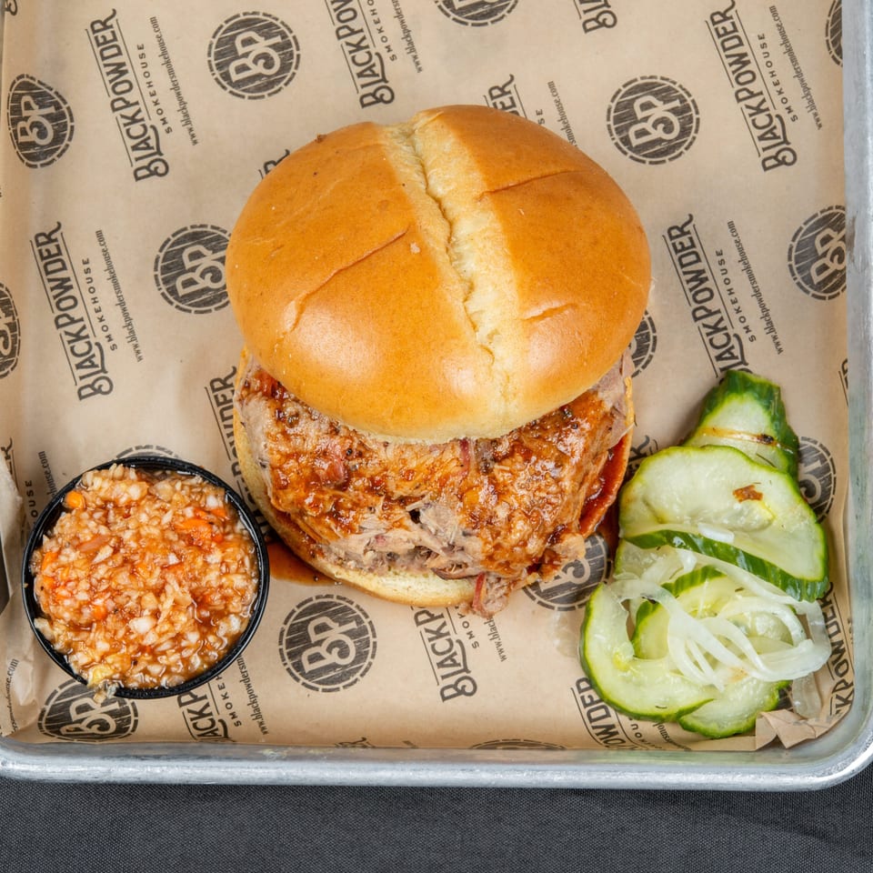 Pulled pork sandwich with barbecue sauce, rice side, and sliced cucumbers and onions on a Black Powder Smokehouse tray.