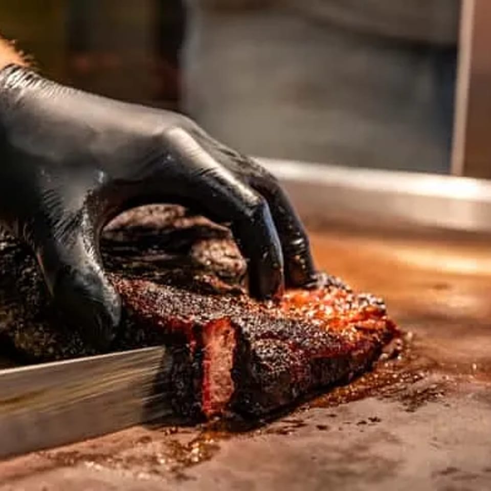 Gloved hand slicing a large, smoked brisket with a knife on a metal surface.