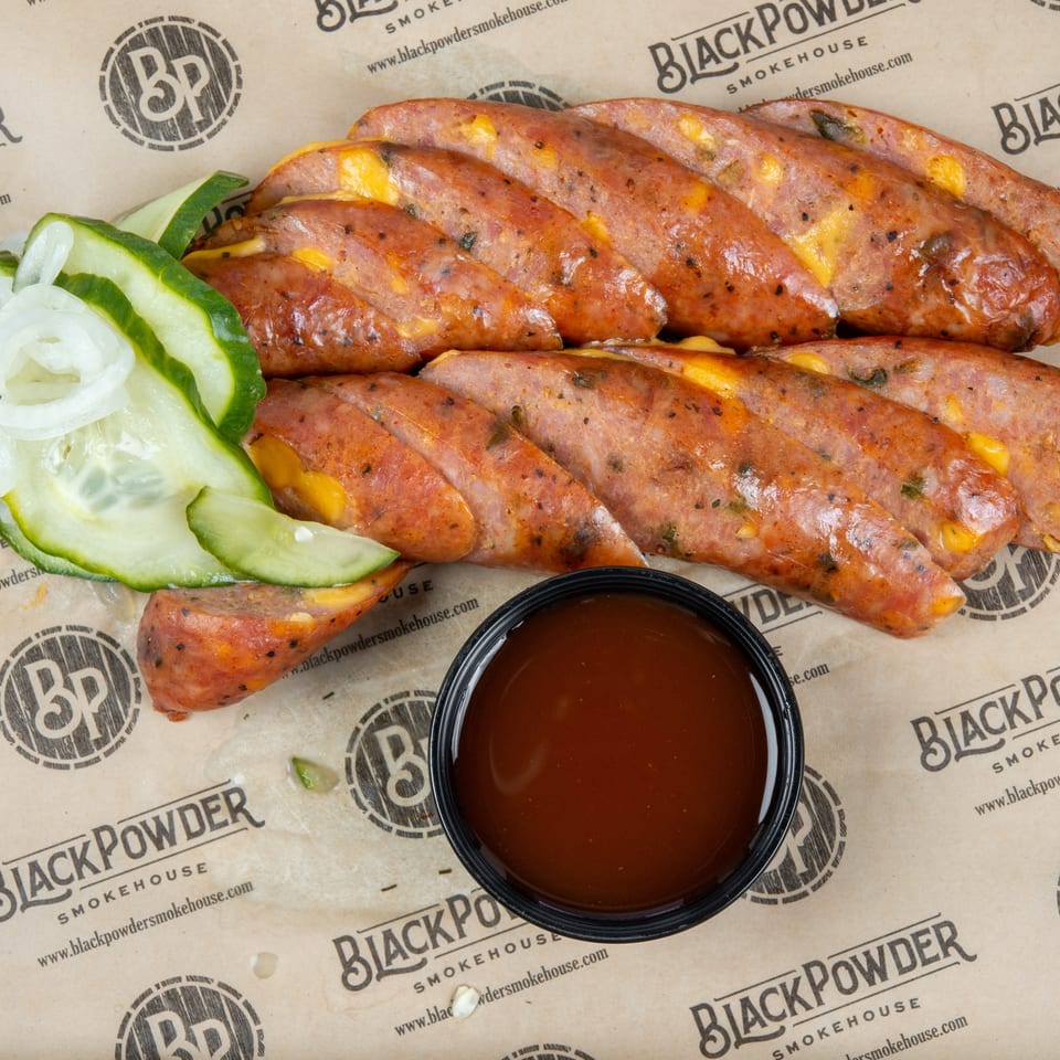 Grilled cheese-and-jalapeño sausages with sliced pickles, onions, and dipping sauce on Black Powder Smokehouse paper.