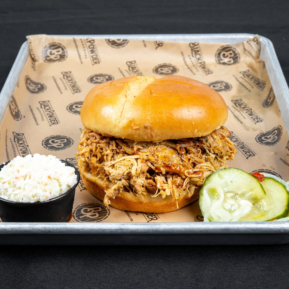 Pulled pork sandwich on a toasted bun served with coleslaw and sliced pickles in a branded metal tray.