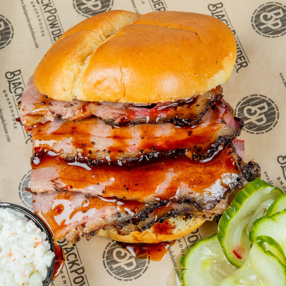 Saucy stacked barbecue sandwich on a bun, served with creamy coleslaw and sliced pickles on Black Powder Smokehouse paper.
