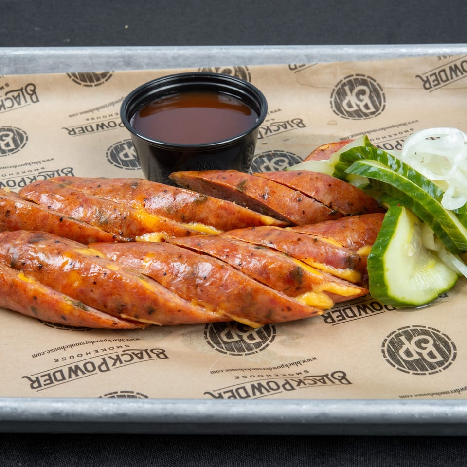 Grilled, cheese-stuffed sausages served with sliced cucumbers, onions, and dipping sauce on branded paper.