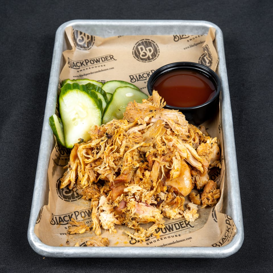 Pulled smoked pork with sliced pickles and barbecue sauce in a Black Powder Smokehouse-lined metal tray.