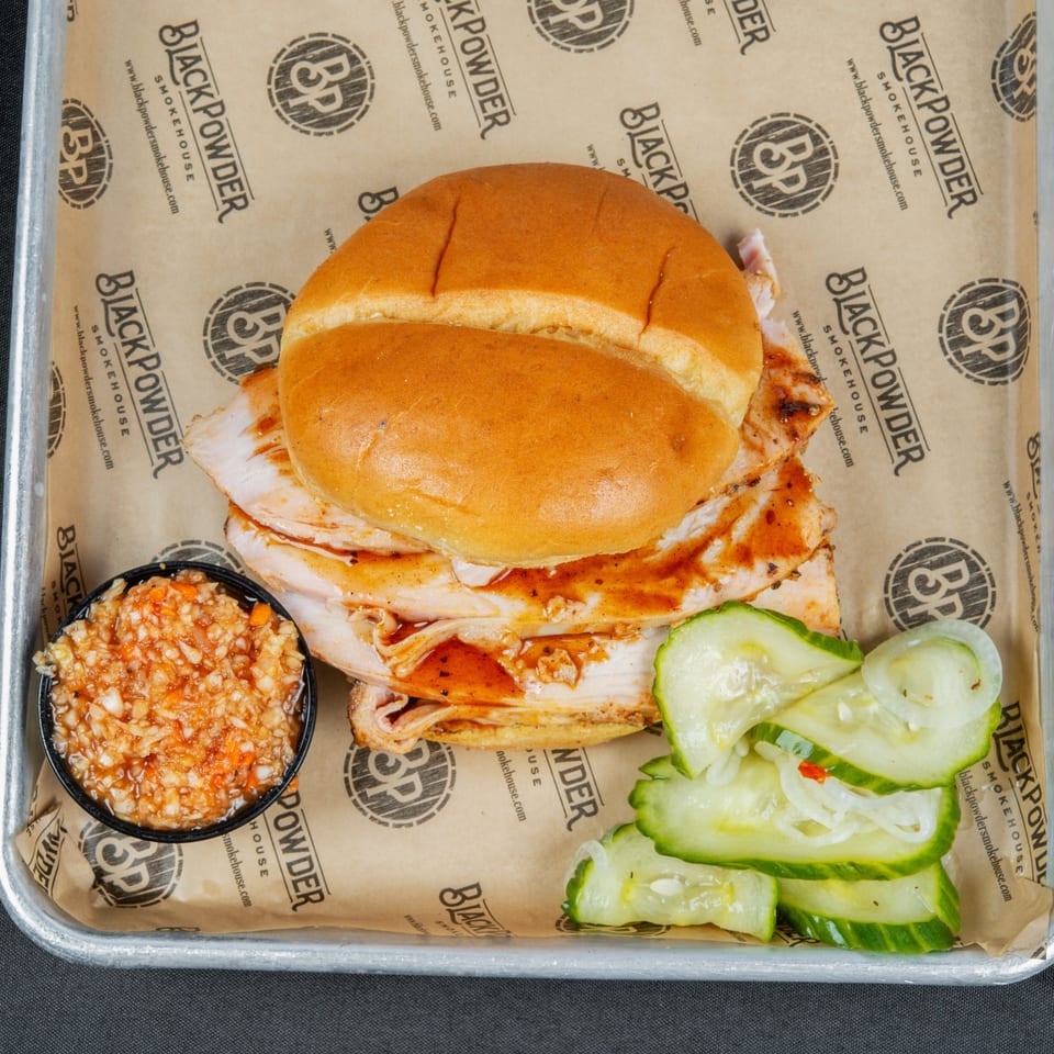 Pulled pork sandwich with barbecue sauce, pickles, and a side of relish on Black Powder Smokehouse paper.