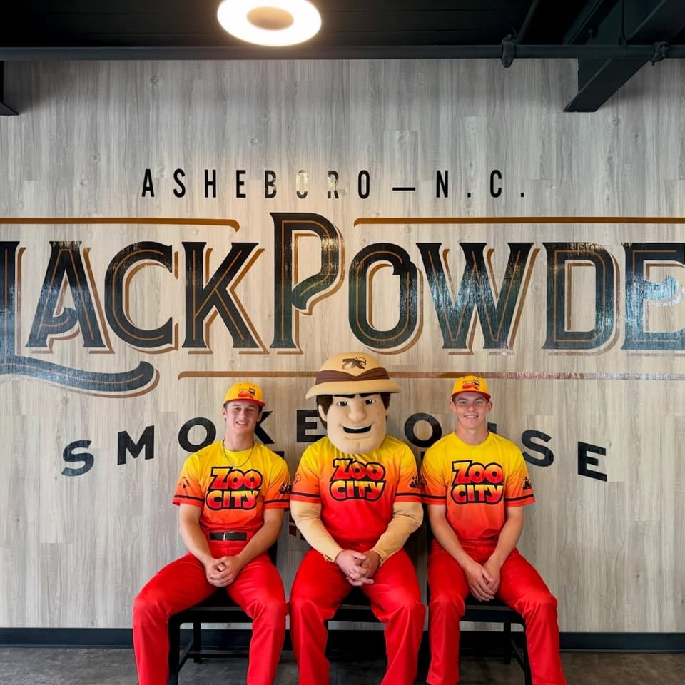 Two men and a mascot in red and yellow outfits sit in front of a "Black Powder Smokehouse" sign.