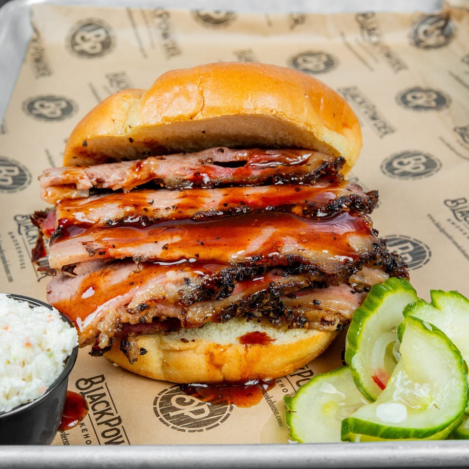 Saucy pulled pork sandwich piled with smoked meat, served with coleslaw and sliced pickles on a restaurant tray.